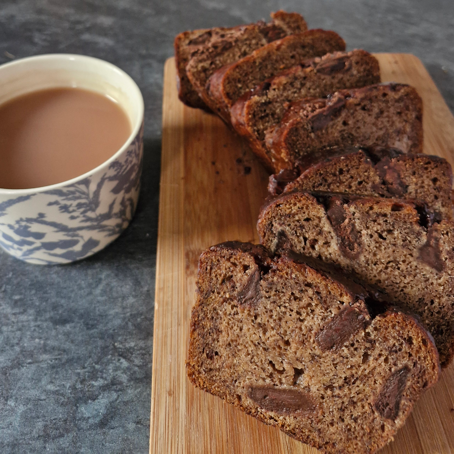 Slices of chocolate chip bread arranged on a wooden cutting board next to a blue and white patterned cup of coffee on a gray stone surface.