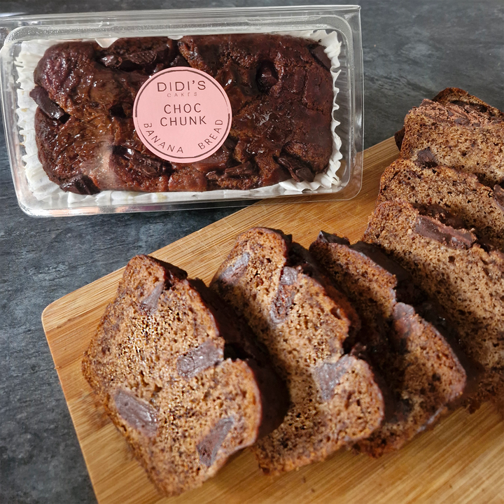 Chocolate chunk banana bread shown as a sliced loaf on a wooden cutting board next to its clear plastic packaging container with a pink circular label reading &