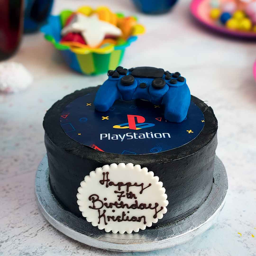 A round birthday cake decorated with a PlayStation logo and a blue playstation controller on top, with a 'Happy 7th Birthday' plaque, set on a table with other birthday treats in the background.