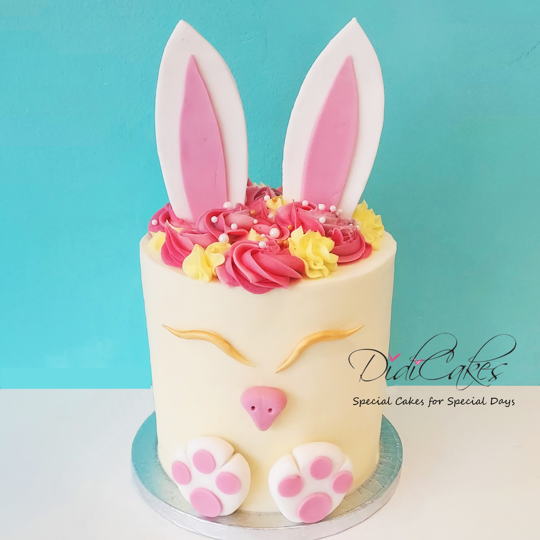 A decorated cake designed to look like a bunny with white frosting and pink details, featuring bunny ears and paws, with yellow and pink frosting flowers on top.